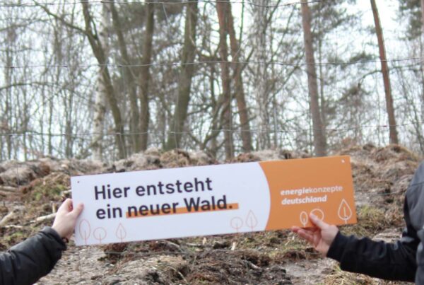 Zwei Menschen vor einem Wald, die eine Schild halten auf dem steht "Hier entsteht ein neuer Wald"