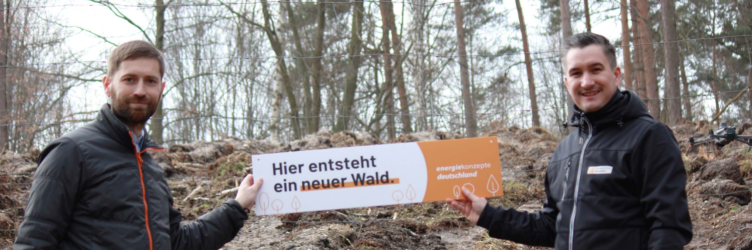 Zwei Menschen vor einem Wald, die eine Schild halten auf dem steht "Hier entsteht ein neuer Wald"