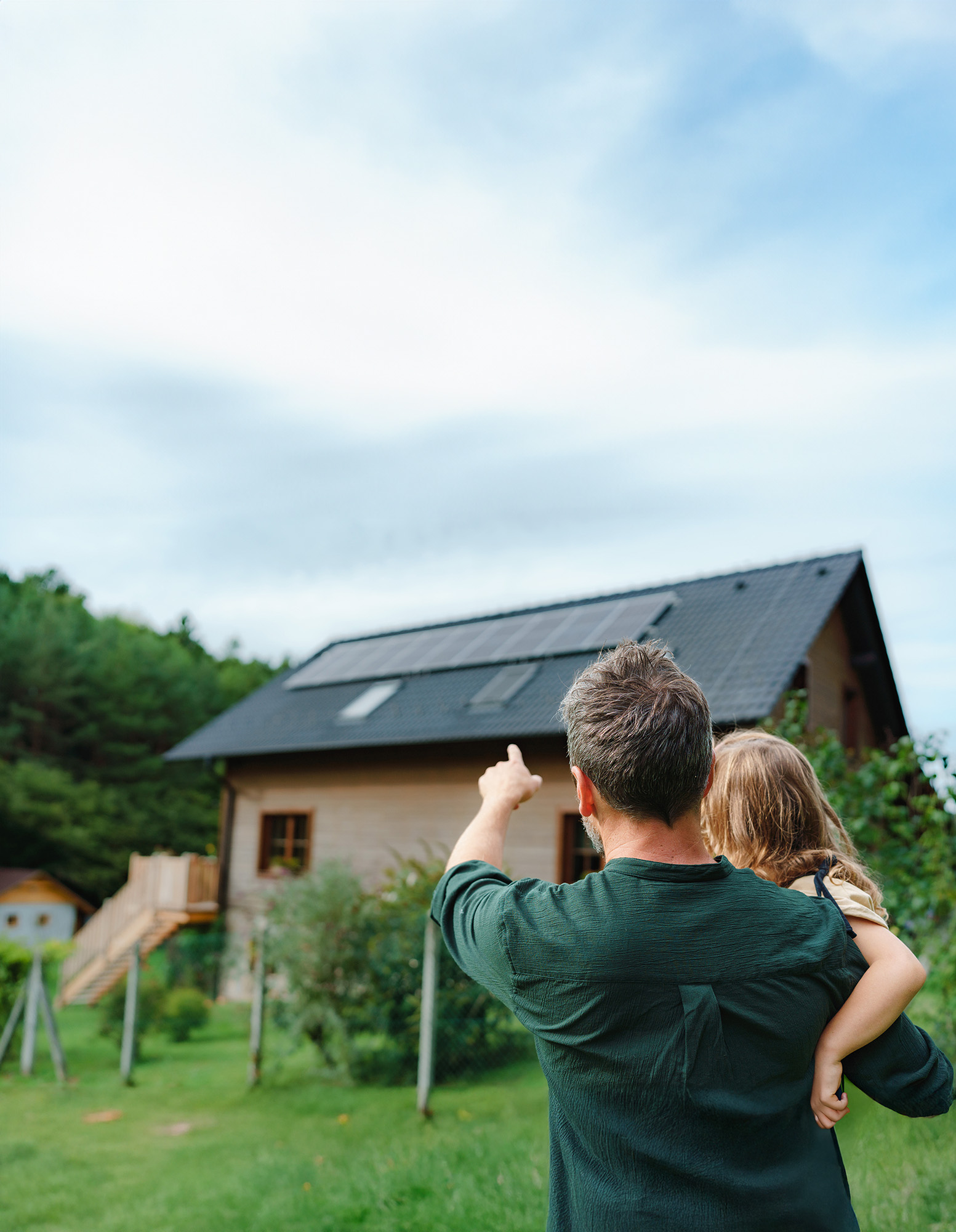 Mann hält Tochter auf dem Arm und zeigt auf ein Haus mit Solaranlage.